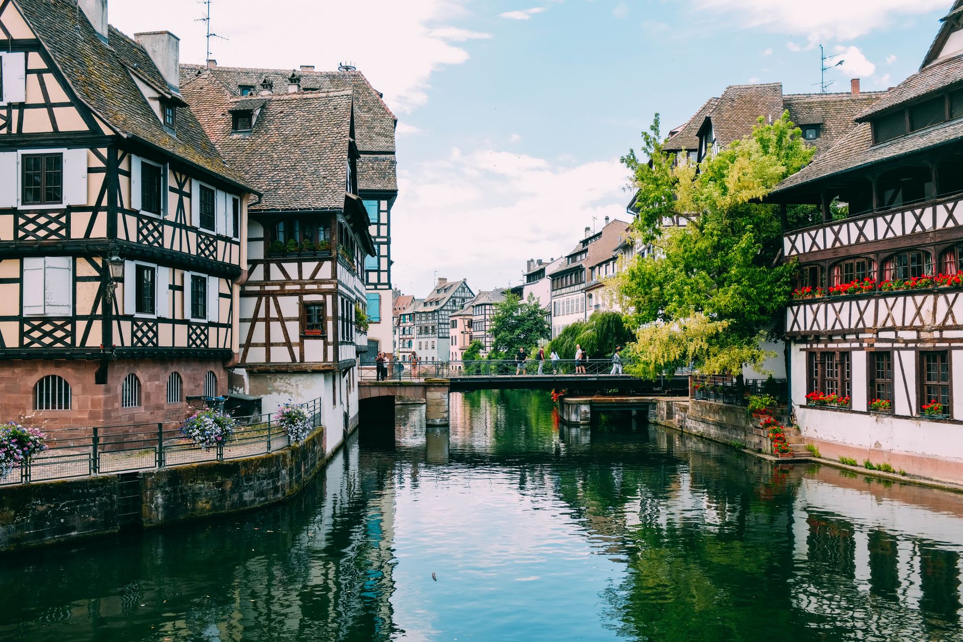 Canal bordé de maisons à colombages fleuries, avec une passerelle où marchent quelques personnes, sous un ciel nuageux.