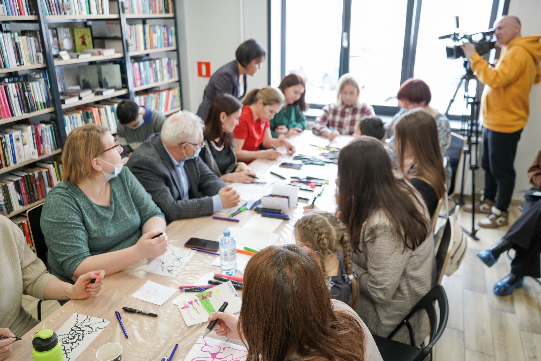 Enfants et adultes dessinent autour d’une table avec des feutres dans une bibliothèque, devant des rayonnages de livres, tandis qu’un cameraman filme.