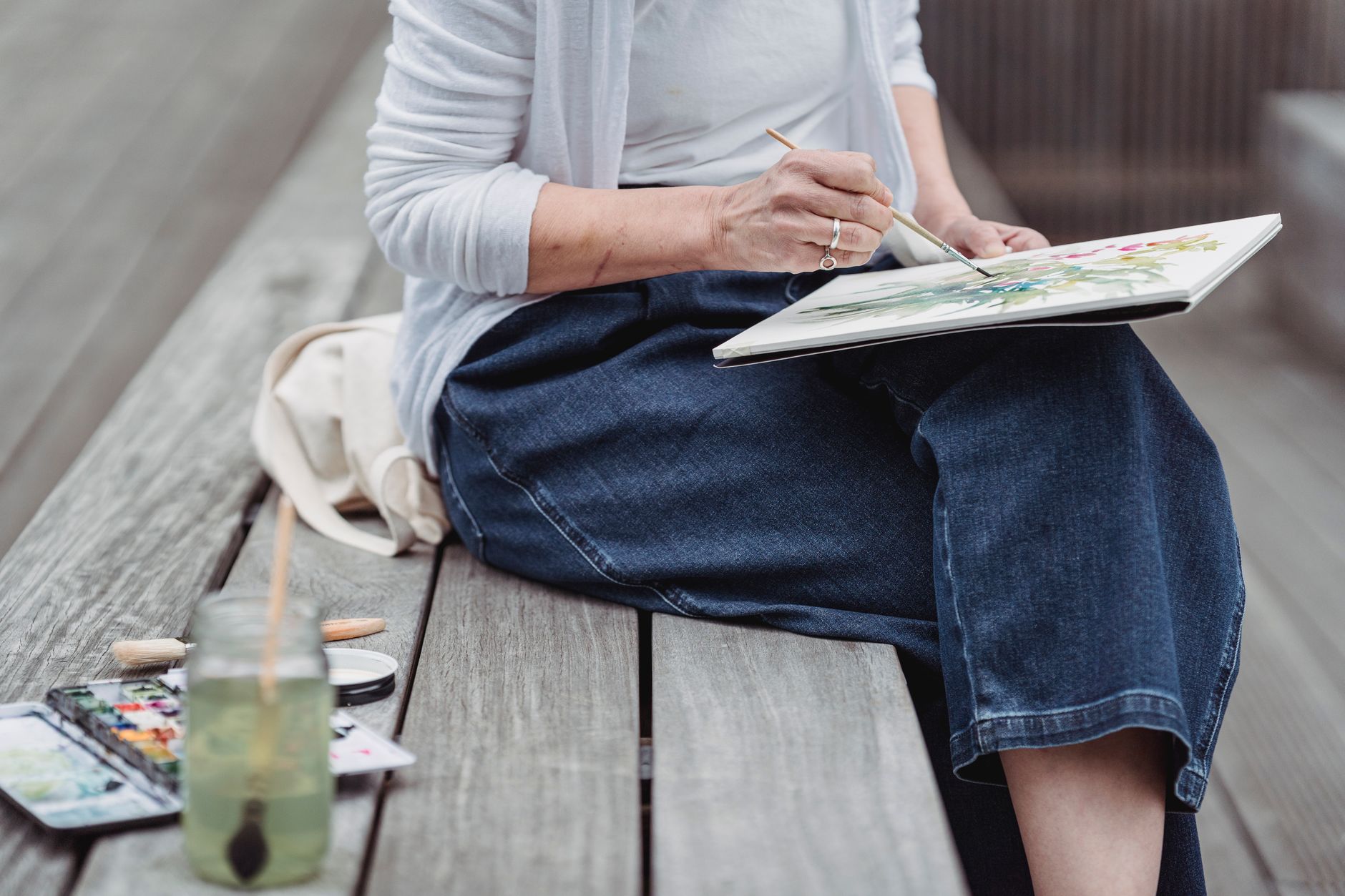 Personne assise sur un banc en bois peignant à l’aquarelle, avec une boîte de peinture et un bocal d’eau posés à côté.