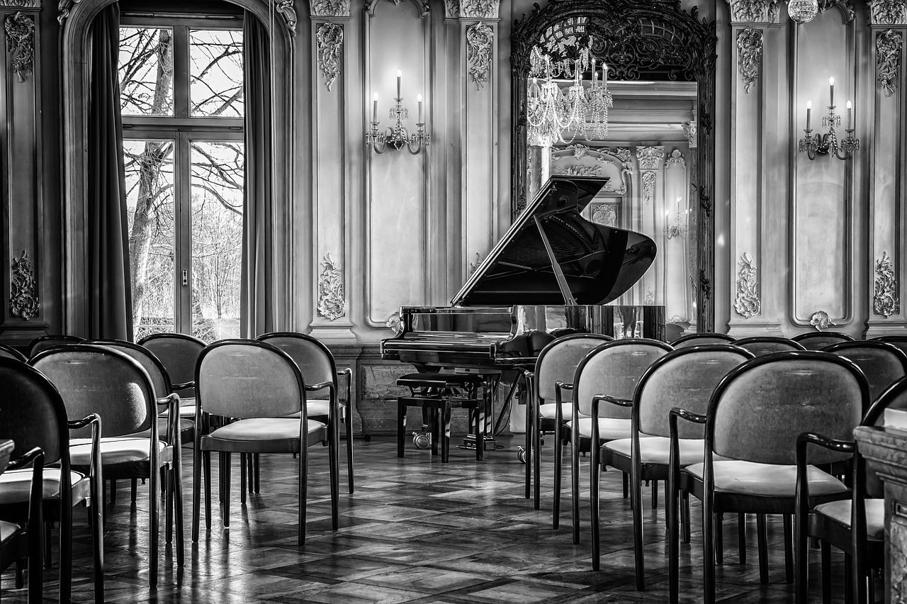 Un salon de musique aux murs moulurés, avec un piano à queue et des rangées de chaises vides, en noir et blanc