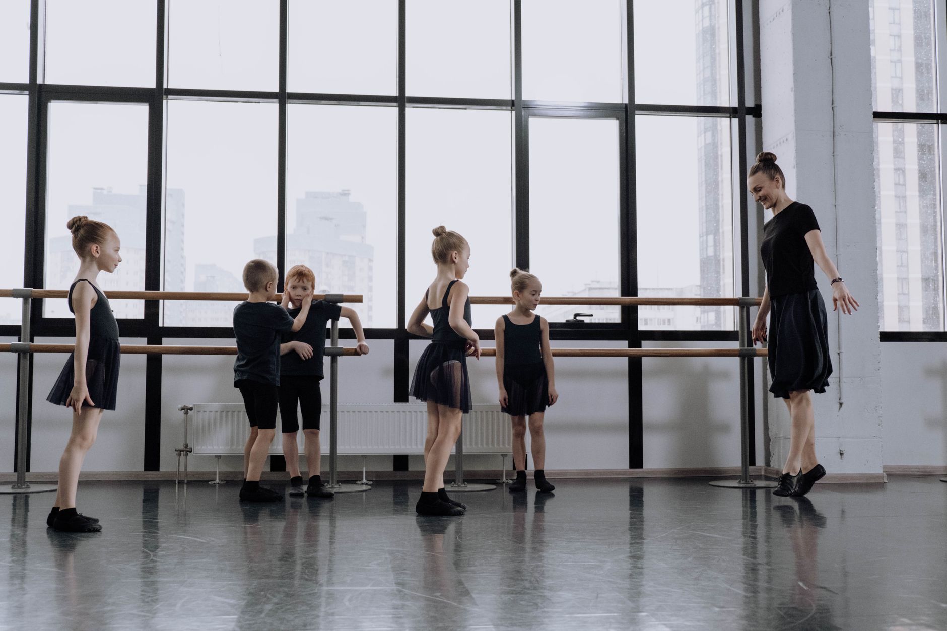 Une professeure et des enfants en tenue sombre dans une salle de danse devant de grandes fenêtres.